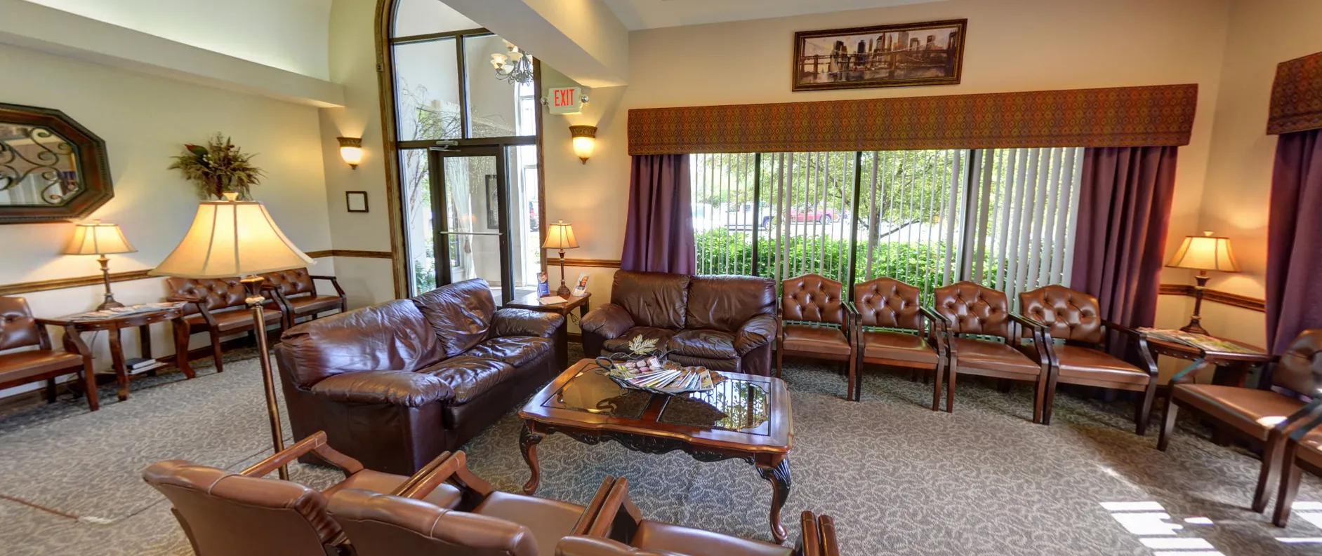 The C. Patrick Gray, DDS waiting room in Flint, MI, featuring leather sofas and armchairs, warm lamp lighting, large windows with purple drapes, and a glass-top coffee table with reading materials.