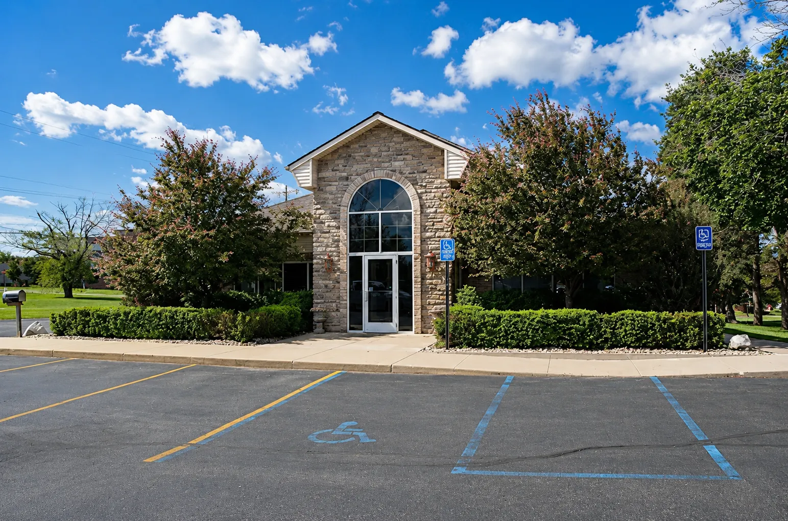 Front entrance of the C. Patrick Gray, DDS. office in Flint, MI, showing a stone brick building with a tall arched window above the main glass doors, flanked by trimmed hedges and trees, with accessible parking spaces visible in the foreground.