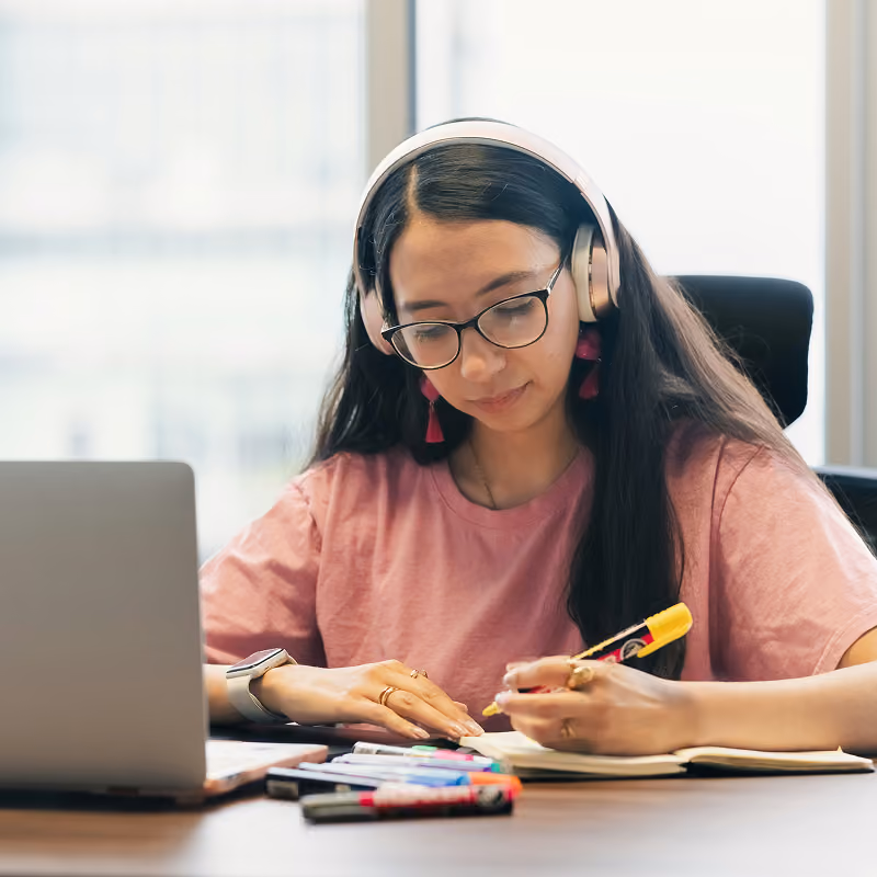 Young woman with long dark hair and glasses wearing headphones, writing in a notebook at a desk with a laptop and colorful markers.