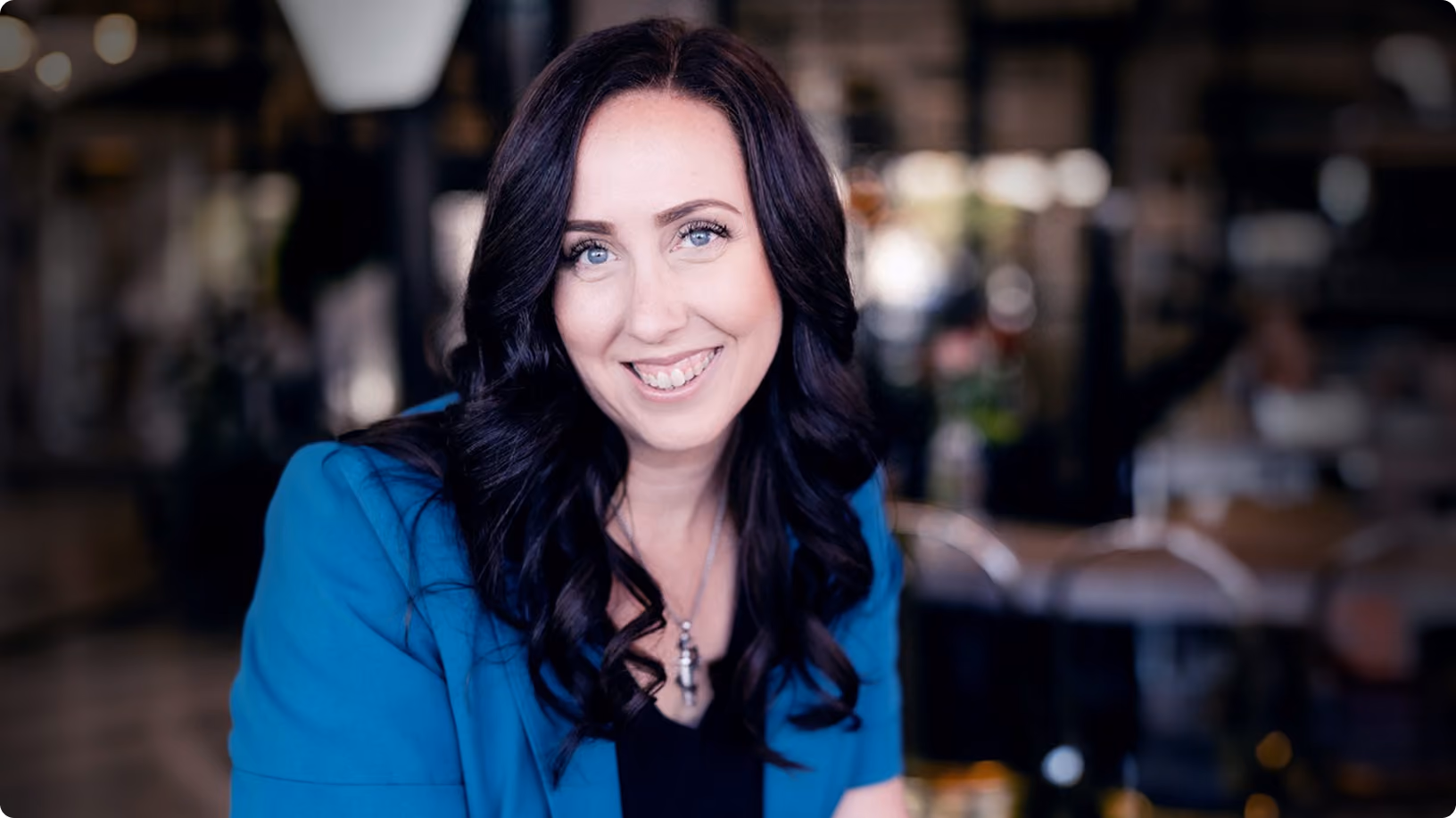 Smiling woman with long dark hair wearing a blue blazer and necklace, sitting indoors with blurred background.