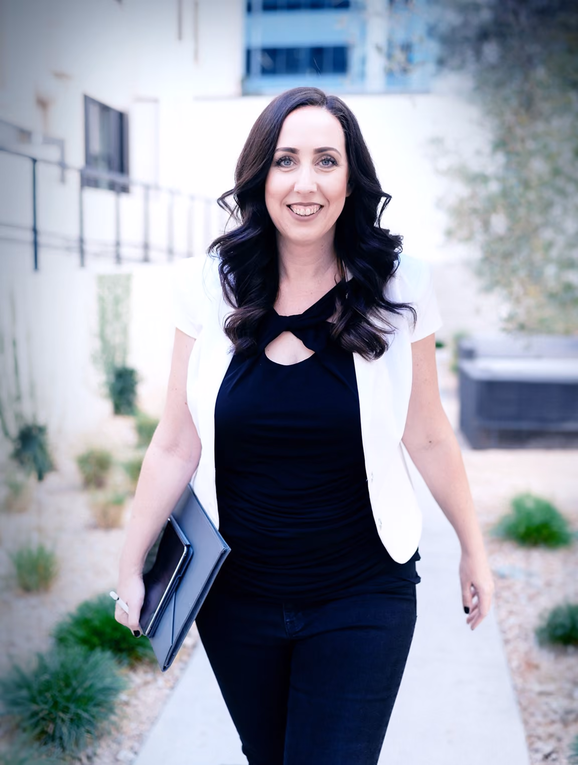 Smiling woman with long dark wavy hair walking outdoors on a paved path between plants, wearing a white short-sleeve jacket over a black top and black pants, holding a folder in one hand.