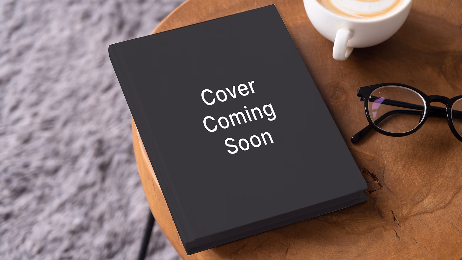 Black book with the white text 'Cover Coming Soon' on the front, placed on a wooden table beside a pair of black-rimmed glasses and a cup of coffee with latte art.
