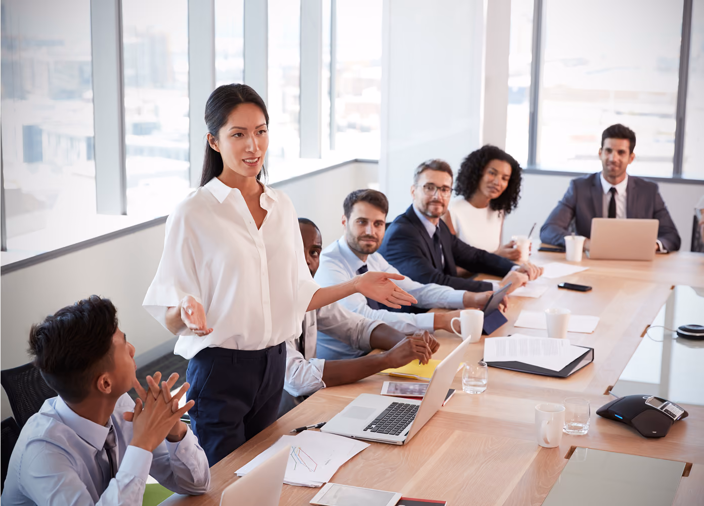 A businesswoman standing and speaking to a diverse group of colleagues seated at a long conference table with laptops, papers, and coffee mugs in a bright office.