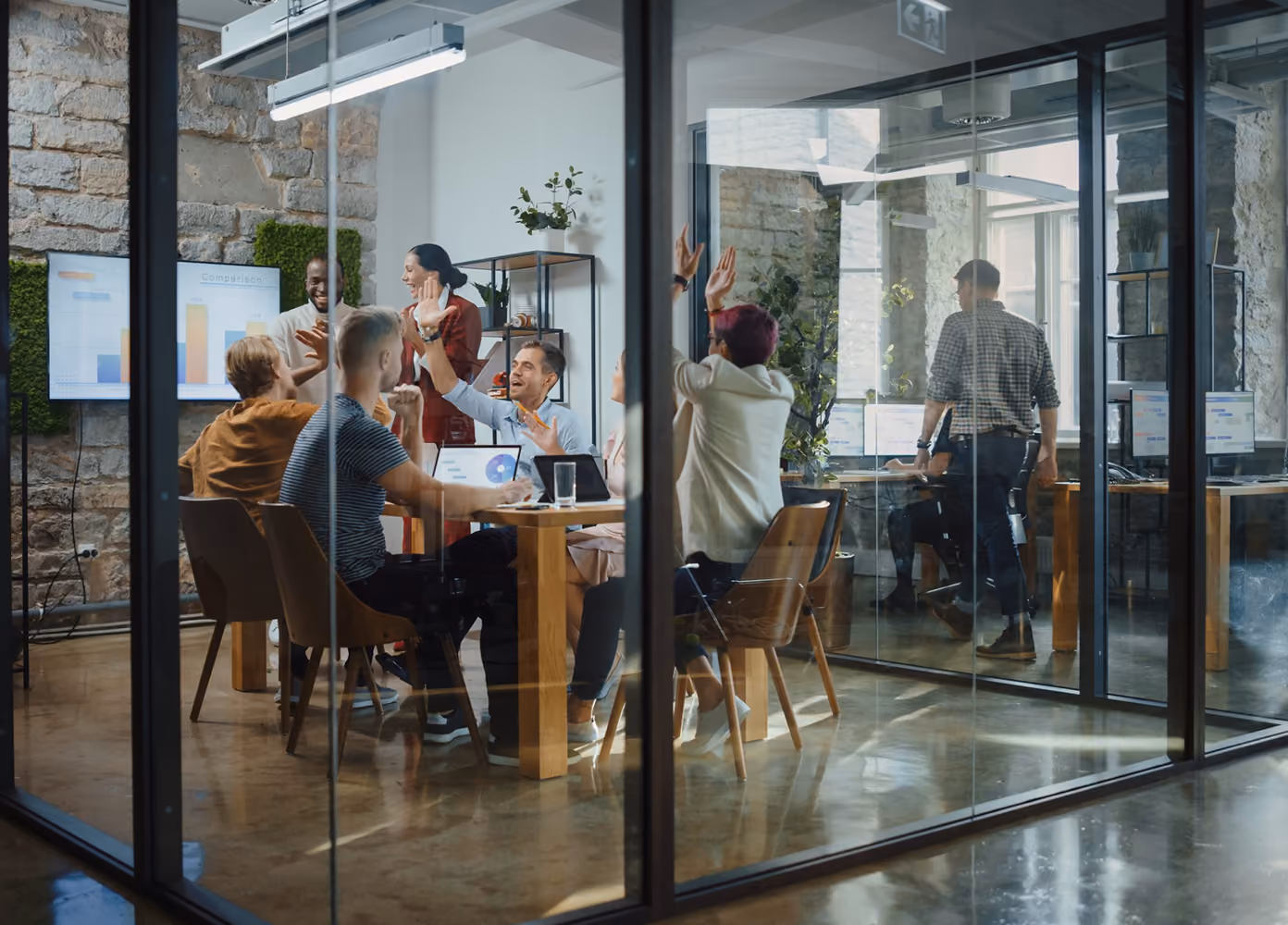A diverse group of professionals enthusiastically high-fiving and collaborating around a wooden conference table in a glass-walled modern office room with stone walls, laptops, and presentation screens displaying graphs.