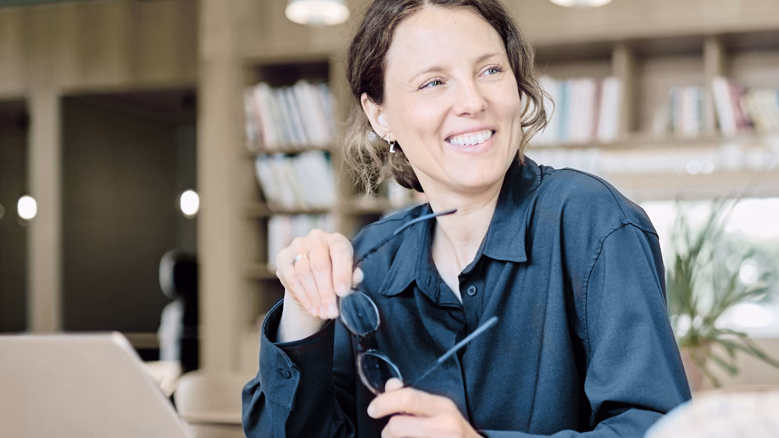 Smiling woman holding eyeglasses in an indoor office setting with bookshelves and plants in the background.