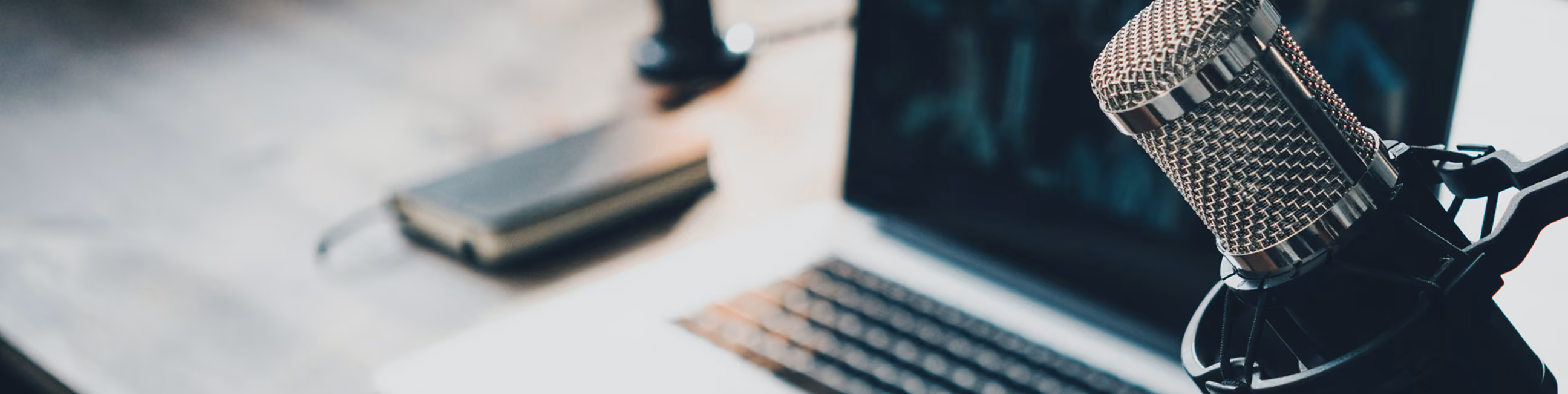 Close-up of a silver microphone on a stand beside a laptop keyboard with a blurred notebook in the background, suggesting a podcast or audio recording setup.