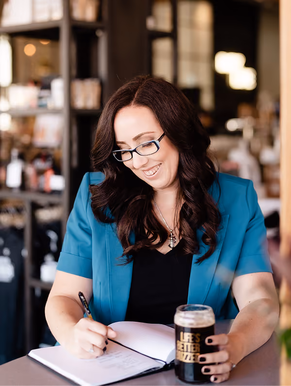 Woman with long dark hair and glasses smiling and writing in a notebook while holding a glass of dark beer at a table in a cozy cafe.