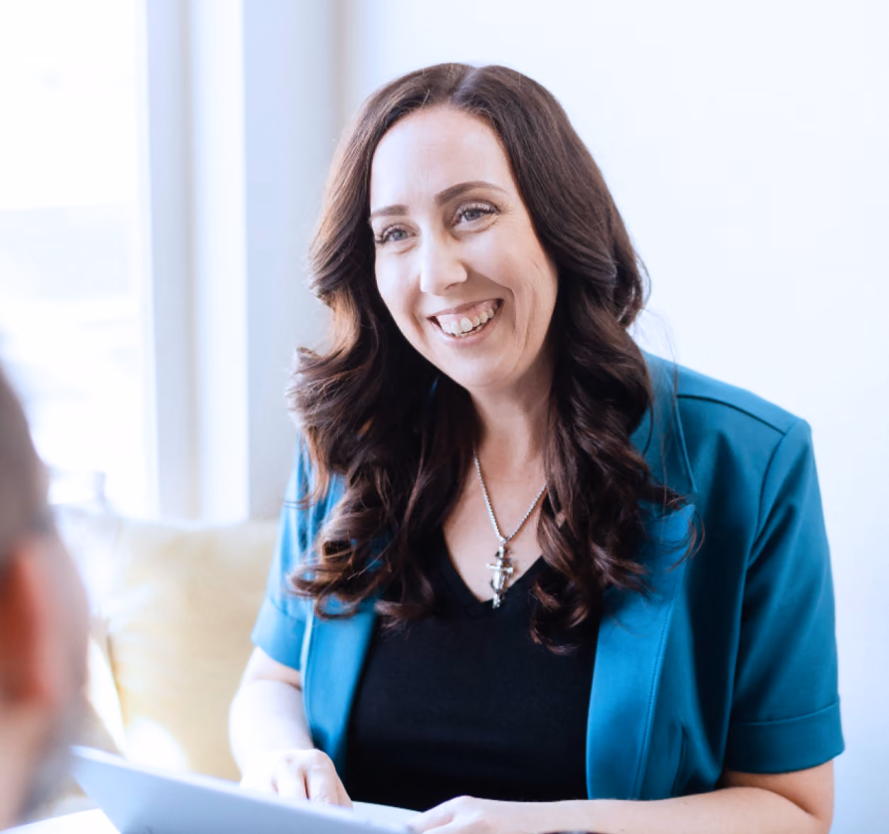 Smiling woman with long curly brown hair wearing a teal jacket and black top, holding a document in a bright room.