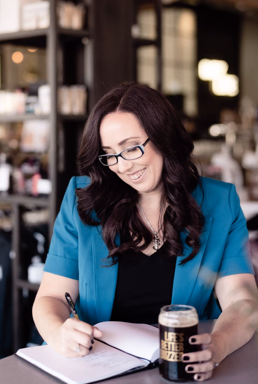 Woman with long dark hair and glasses wearing a teal blazer, smiling while writing in a notebook and holding a glass of dark beer with the text 'LIFE'S BETTER BUZZED'.