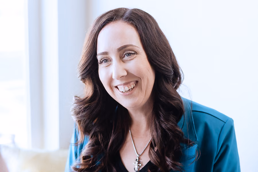 Smiling woman with long dark brown curly hair wearing a teal blazer and a necklace with an anchor pendant in a bright indoor setting.