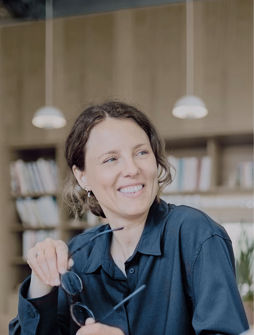 Smiling woman with curly hair wearing a dark blue shirt, holding black-rimmed glasses, sitting at a table with a laptop and an open book, with a blurred bookshelf background.