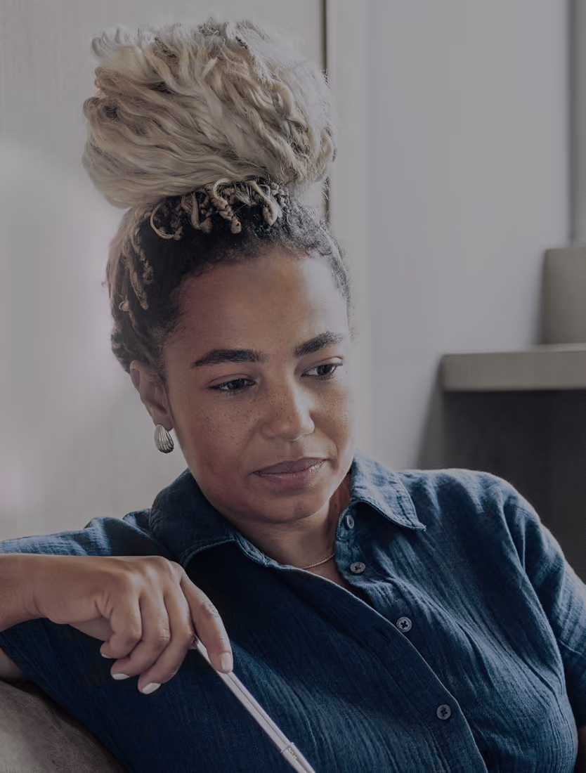 Woman with braided hair and hoop earrings attentively listening while holding a pen and sitting on a couch indoors.