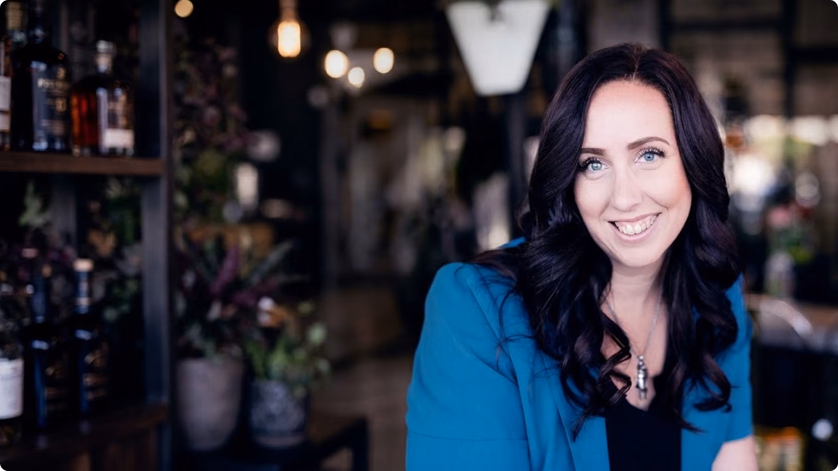 Smiling woman with long dark curly hair wearing a blue blazer and a necklace, sitting indoors with blurred shelves with bottles and plants.