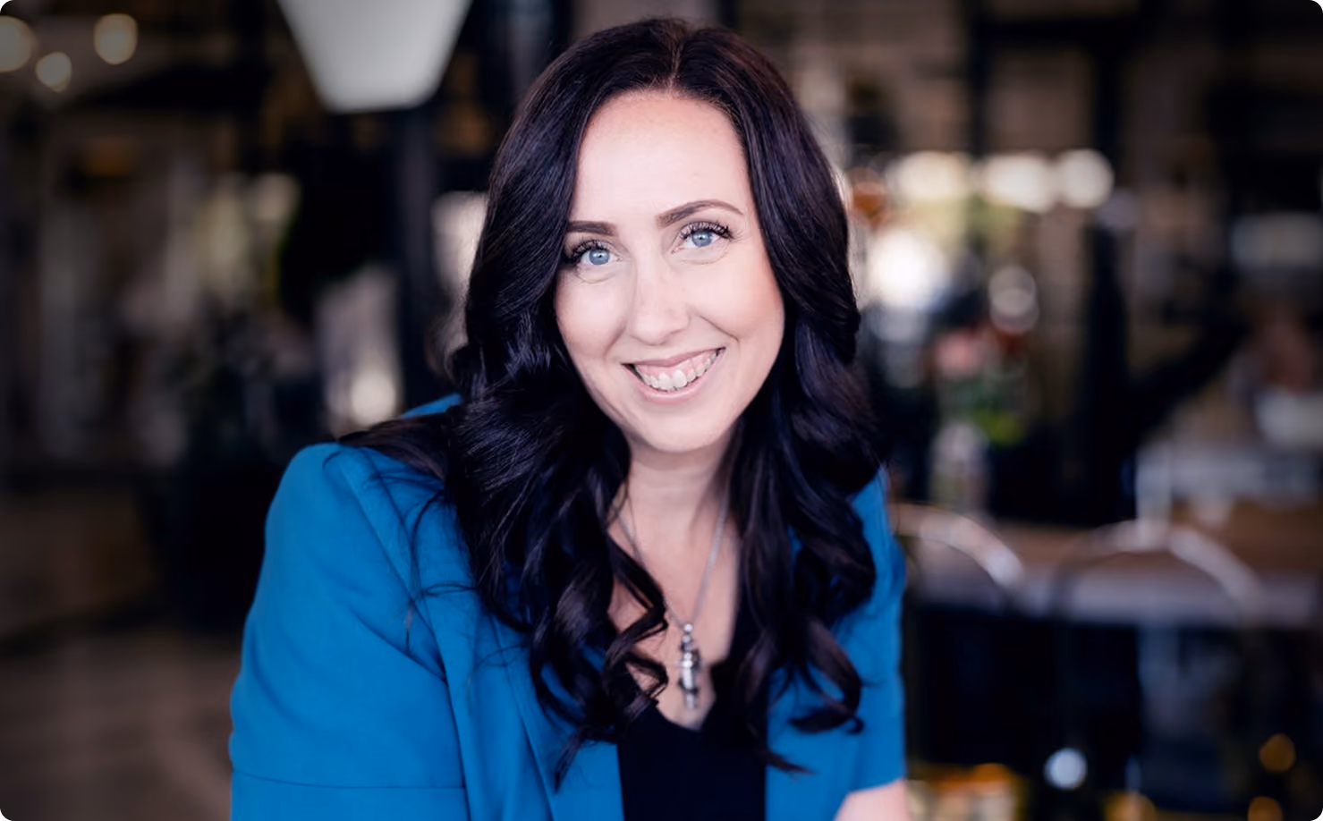 Smiling woman with long dark curly hair wearing a blue blazer over a black top, sitting in a softly blurred indoor setting with warm lighting.