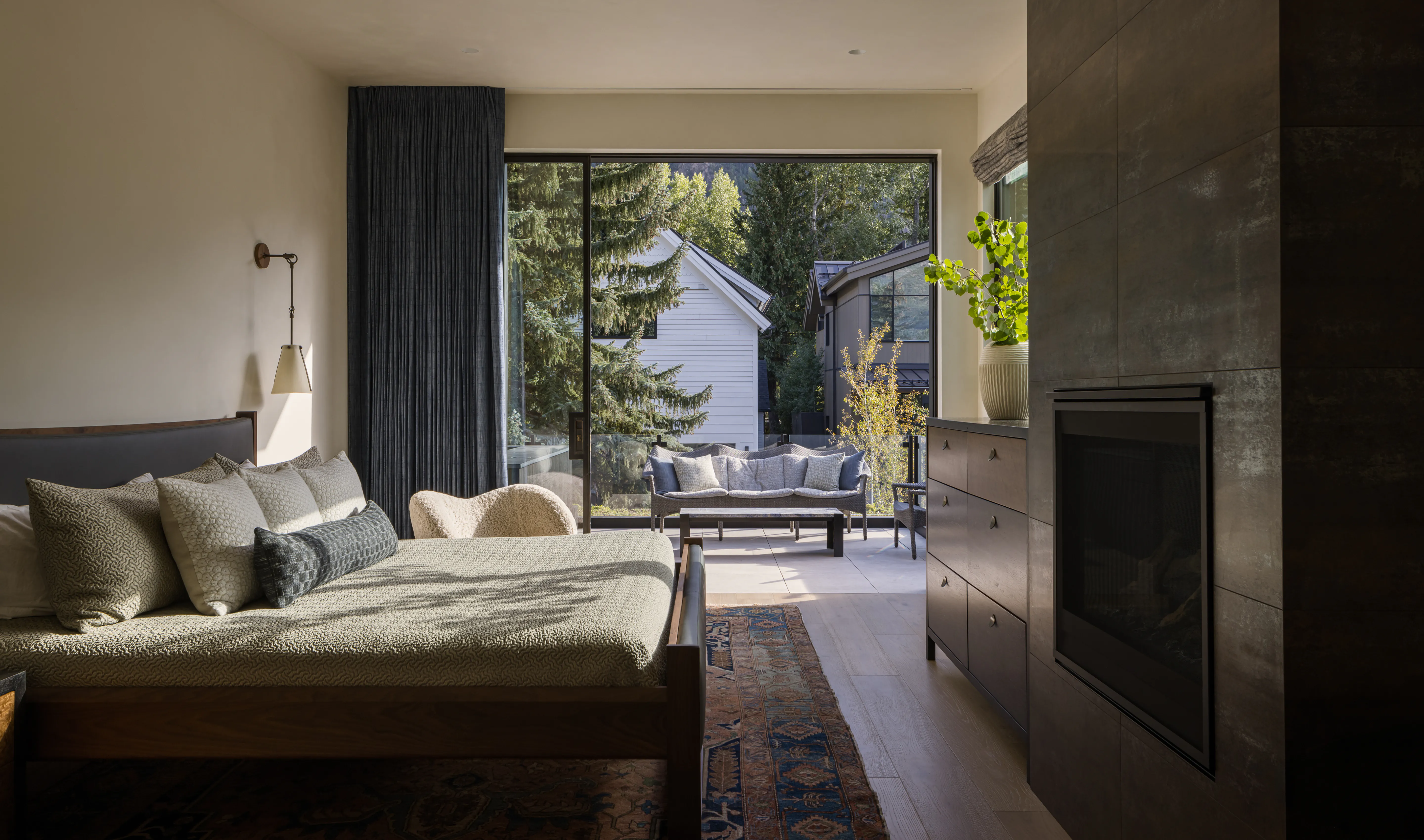 Modern bedroom with large bed, armchair, fireplace, and sliding glass door leading to outdoor seating.