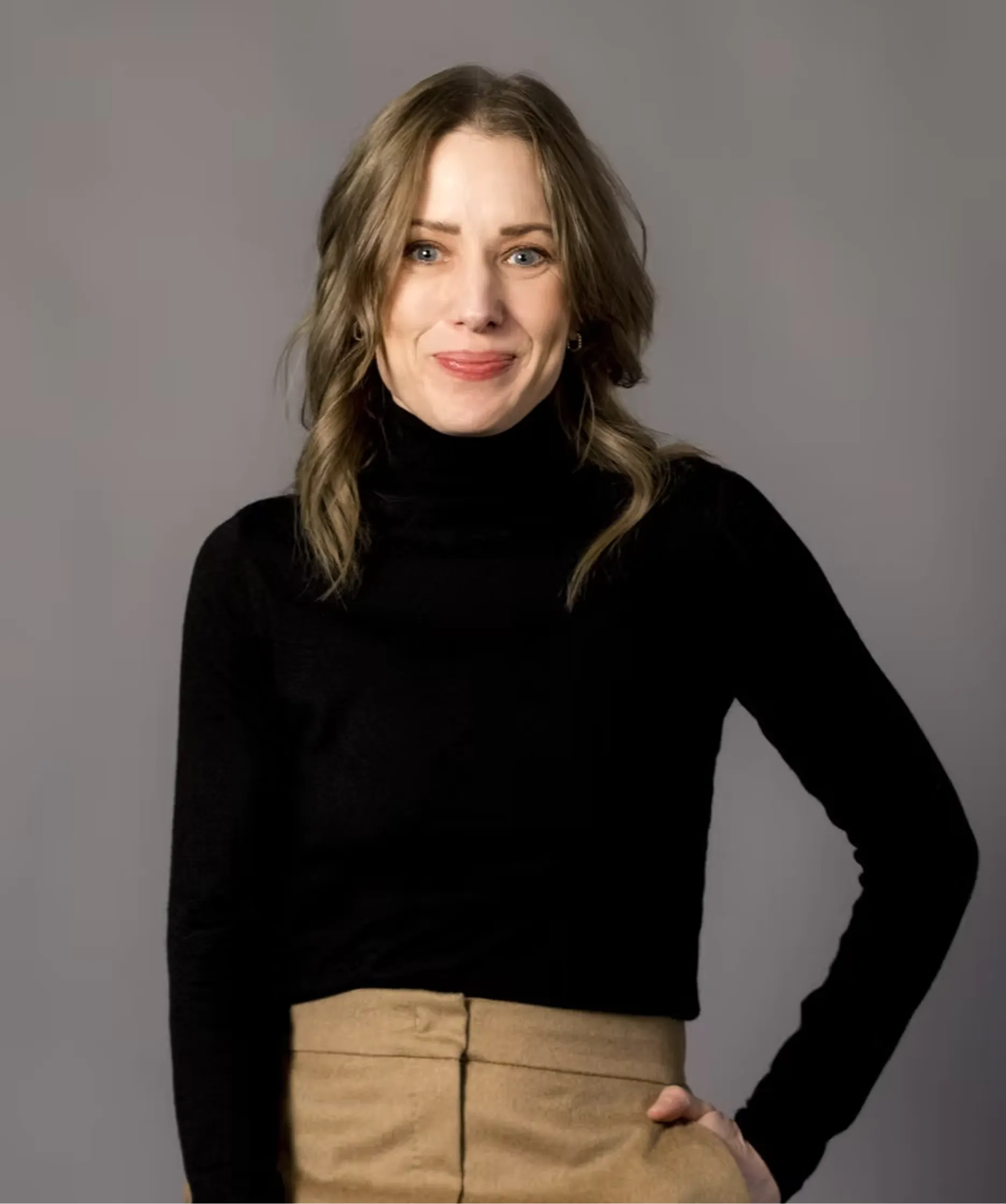 Smiling woman with wavy hair wearing a black turtleneck and tan pants against a gray background.