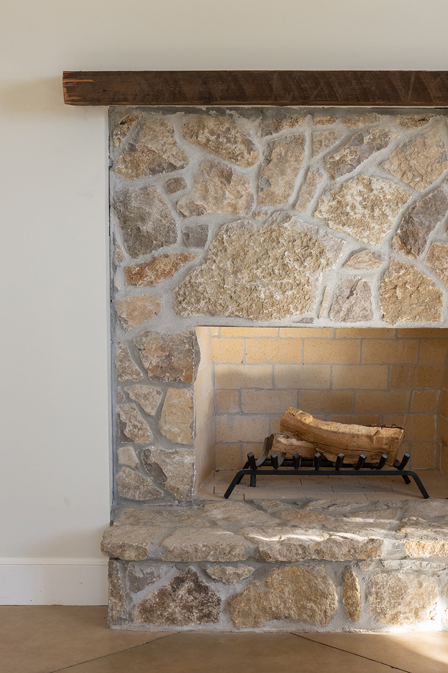 Stone fireplace with light beige and brown rocks, empty inside with a black metal grate holding a single log, topped with a wooden mantel.