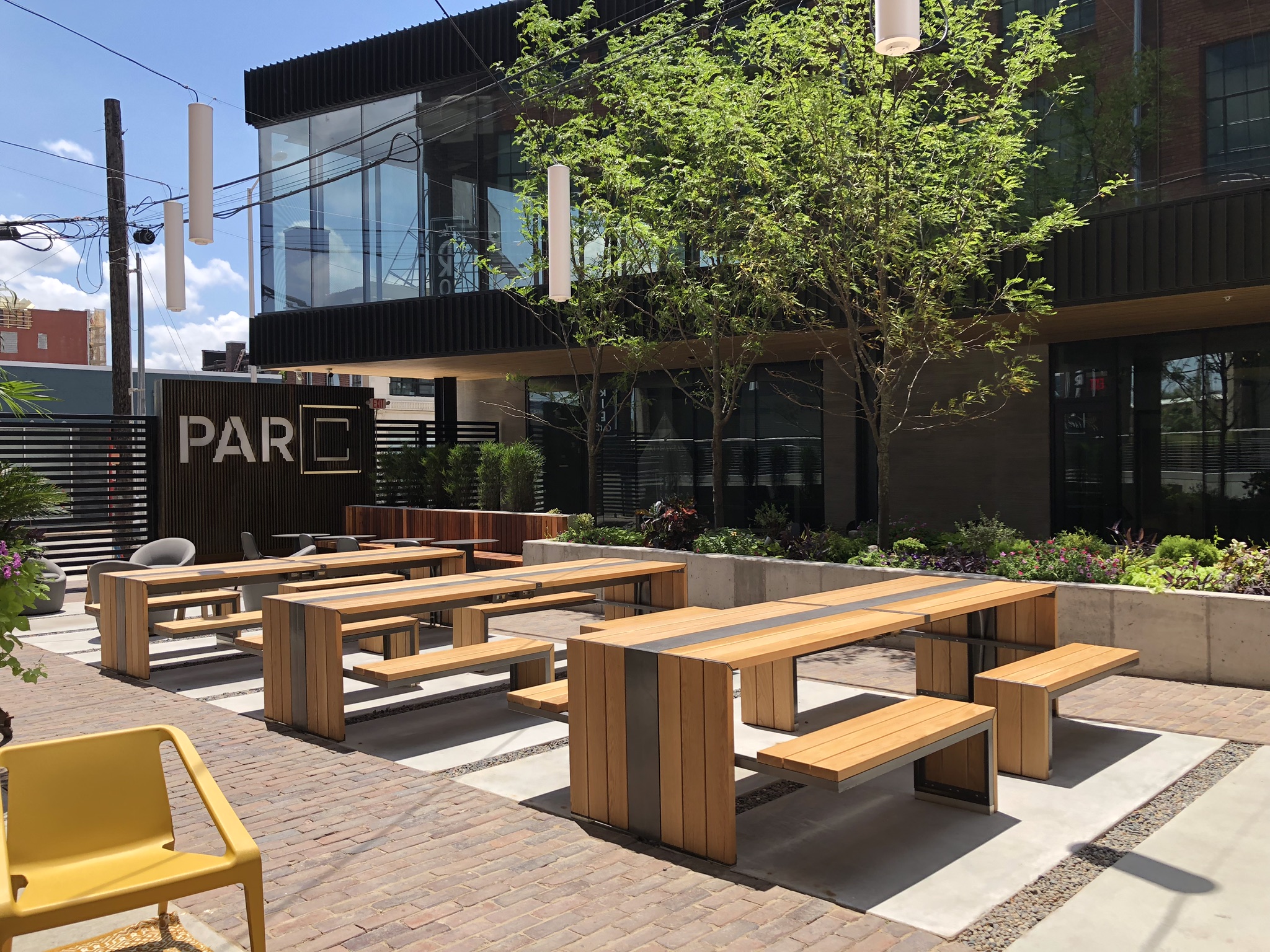 **Alt-text:** Contemporary outdoor dining terrace with custom wood picnic tables and benches, concrete planters, and lush landscaping in front of a modern black-and-brick building. White pendant lights hang overhead in the sunny courtyard.