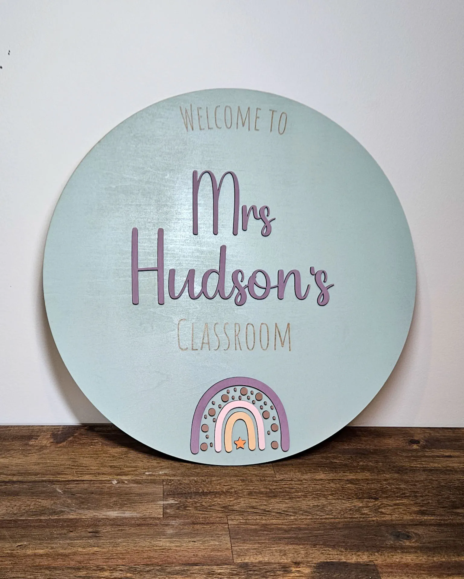 Round pale blue classroom door sign reading Welcome to Mrs Hudson's classroom, propped against a wall