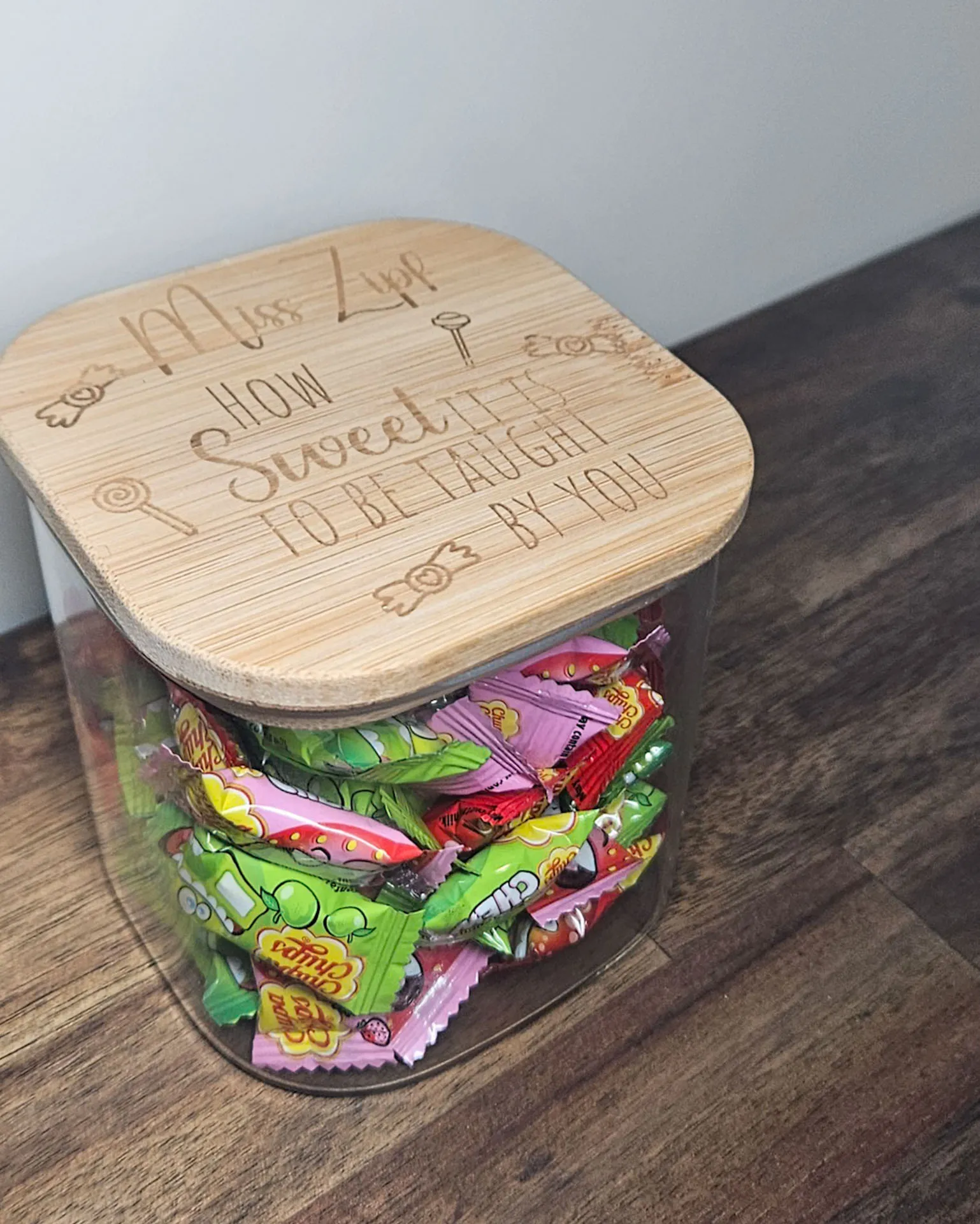 Glass lolly jar filled with wrapped lollies, topped with an engraved bamboo lid reading Mrs Zyd, how sweet it is to be taught by you