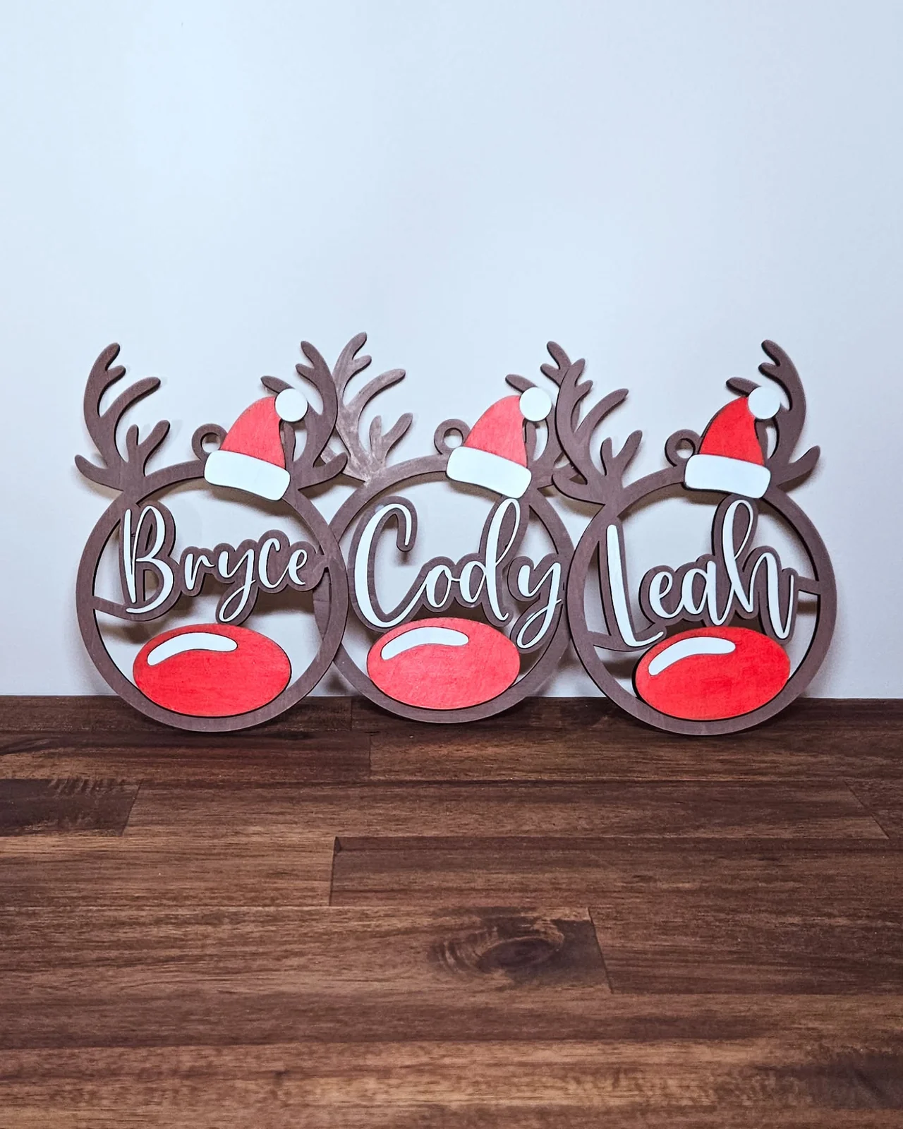 Three cute wooden Christmas ornaments in a reindeer shape with a big red nose, each one shows a name Bryce, Cody and Leah.