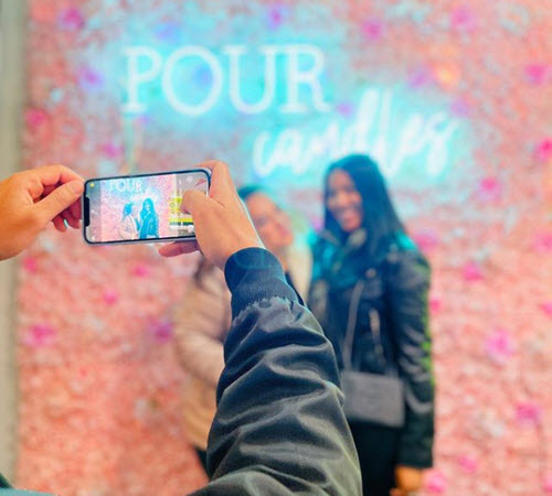 Friend photographing a guest in front of the Pour Candles neon flower wall at Wick & Pour