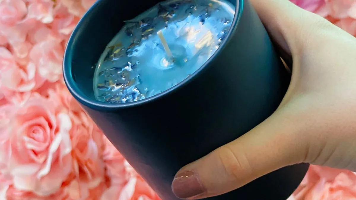 Hand holding a black candle with blue wax and dried petals against pink roses at Wick & Pour
