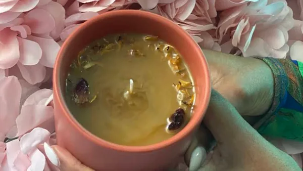 Hand holding a pink candle with golden wax and dried flowers against a floral backdrop