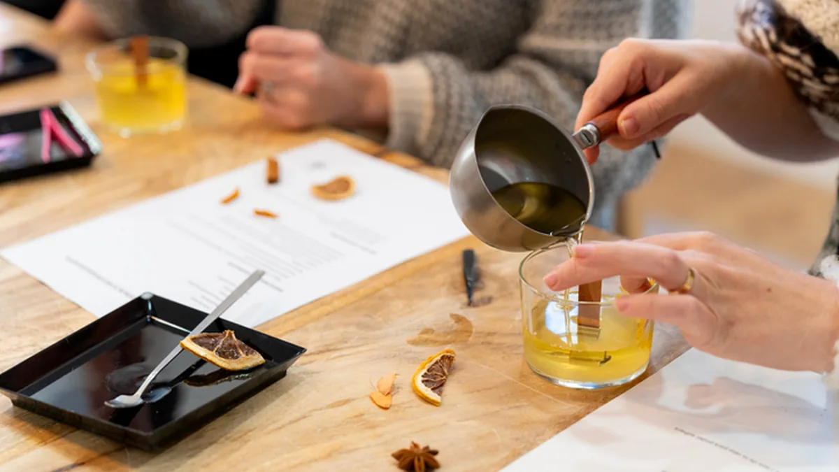 Pouring fragrance oil into a beaker beside dried orange slices and star anise at a candle making class