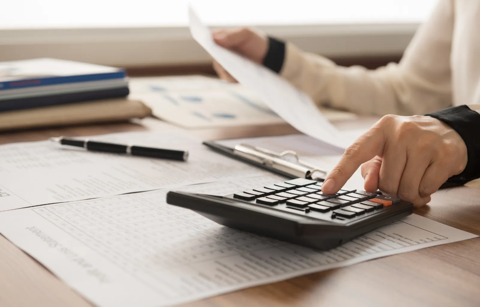 Person using a calculator on a desk covered with financial documents, holding a paper in one hand and pressing calculator buttons with the other, with a pen and folders in the background.