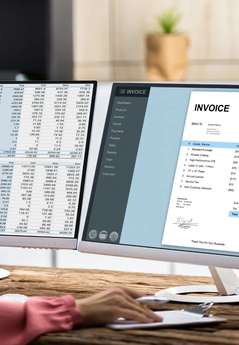 Two computer monitors on a wooden desk showing financial data: the left screen displays spreadsheet tables with numbers by months, the right screen shows an invoicing software interface and a detailed invoice listing various services and prices, with a blurred hand holding a pen over a clipboard in the foreground.