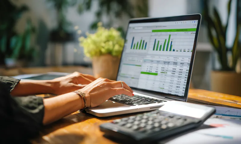 Person typing on a laptop displaying financial data and bar charts, with a calculator and papers on a wooden desk, and a potted plant in the background.