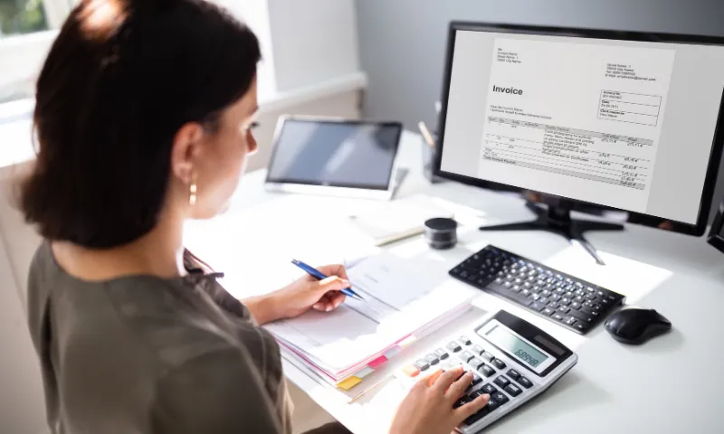 A woman sitting at a desk using a calculator and writing in a notebook with an invoice displayed on a computer monitor in front of her.
