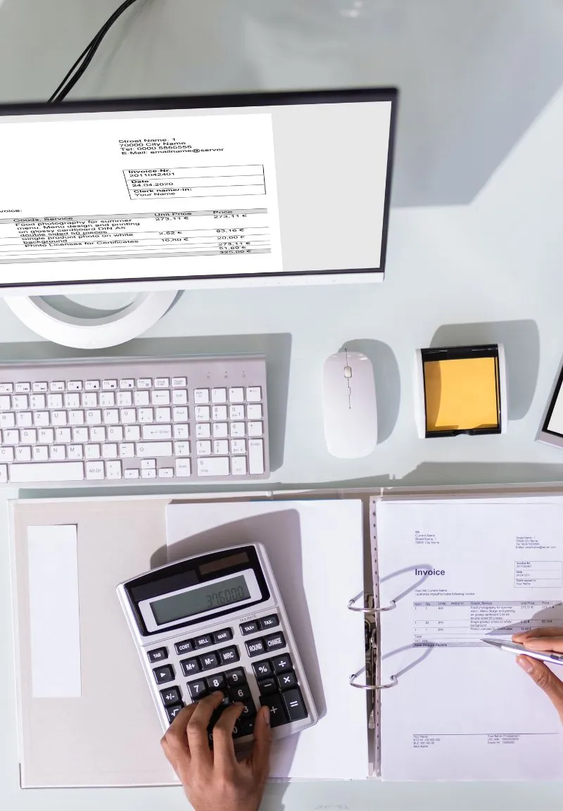 Overhead view of a workspace with a computer monitor displaying an invoice, a white keyboard, a white mouse, a container with yellow sticky notes, and a binder holding an invoice sheet. A hand is pressing calculator buttons, and another hand is pointing at the invoice with a pen.
