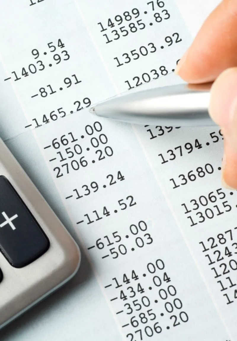 Close-up of a hand holding a silver pen pointing at a financial statement or bank passbook with printed negative and positive numbers, next to a calculator with a plus key visible.