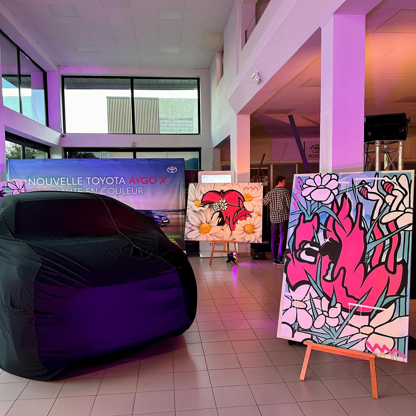 Hall de la concession Toyota éclairé en rose, avec deux toiles de Hugo W2CJ et la voiture encore sous sa housse