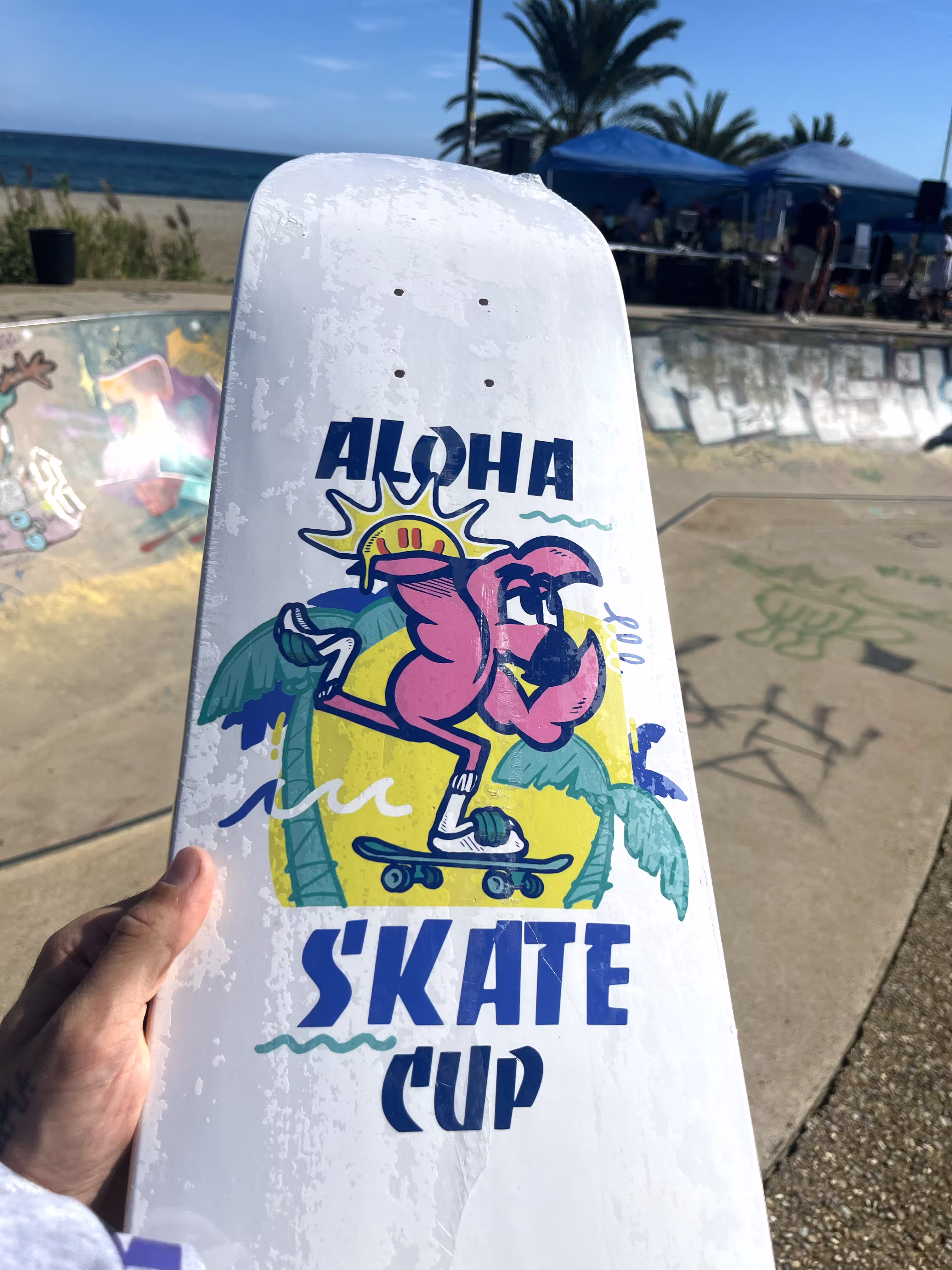 Planche de skate blanche à l'effigie de l'Aloha Skate Cup : flamant rose, soleil et palmiers