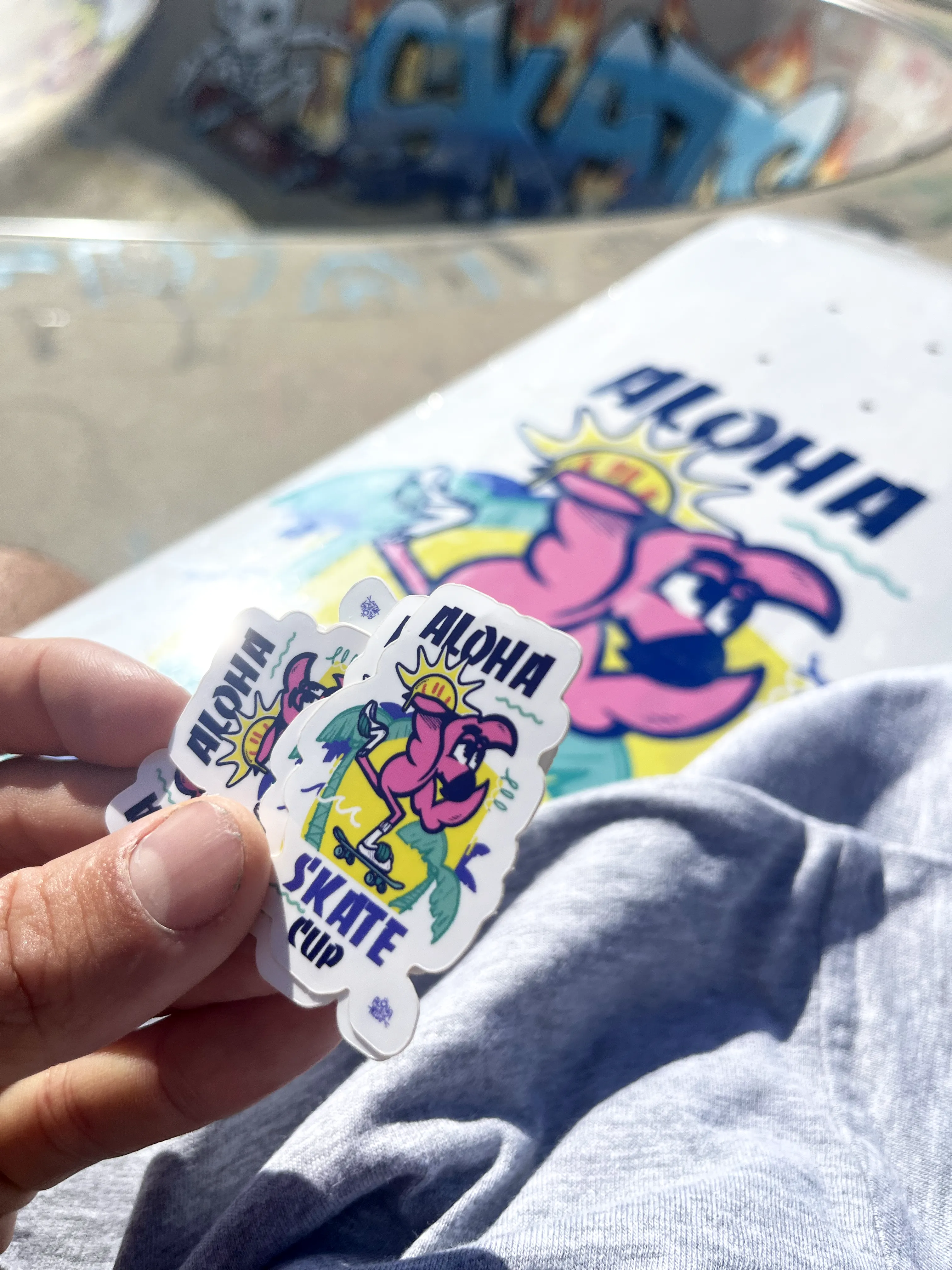 Stickers et planche de l'Aloha Skate Cup au logo dessiné par Hugo W2CJ, tenus devant le bowl