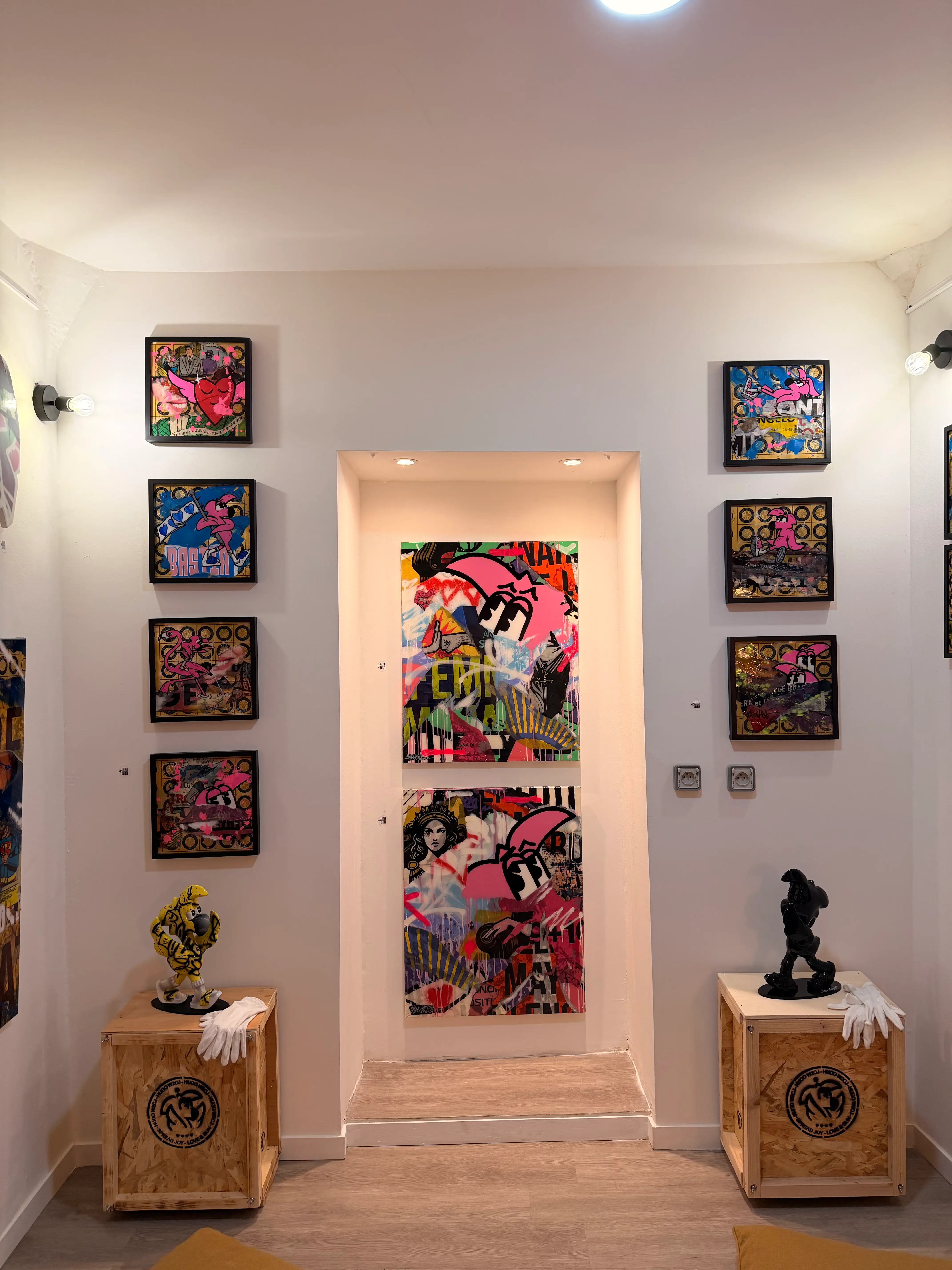 Vue d'ensemble de l'exposition Street Therapy : toiles carrées encadrées autour d'une niche, deux sculptures de Roger le Flamant Rose sur des caisses en bois
