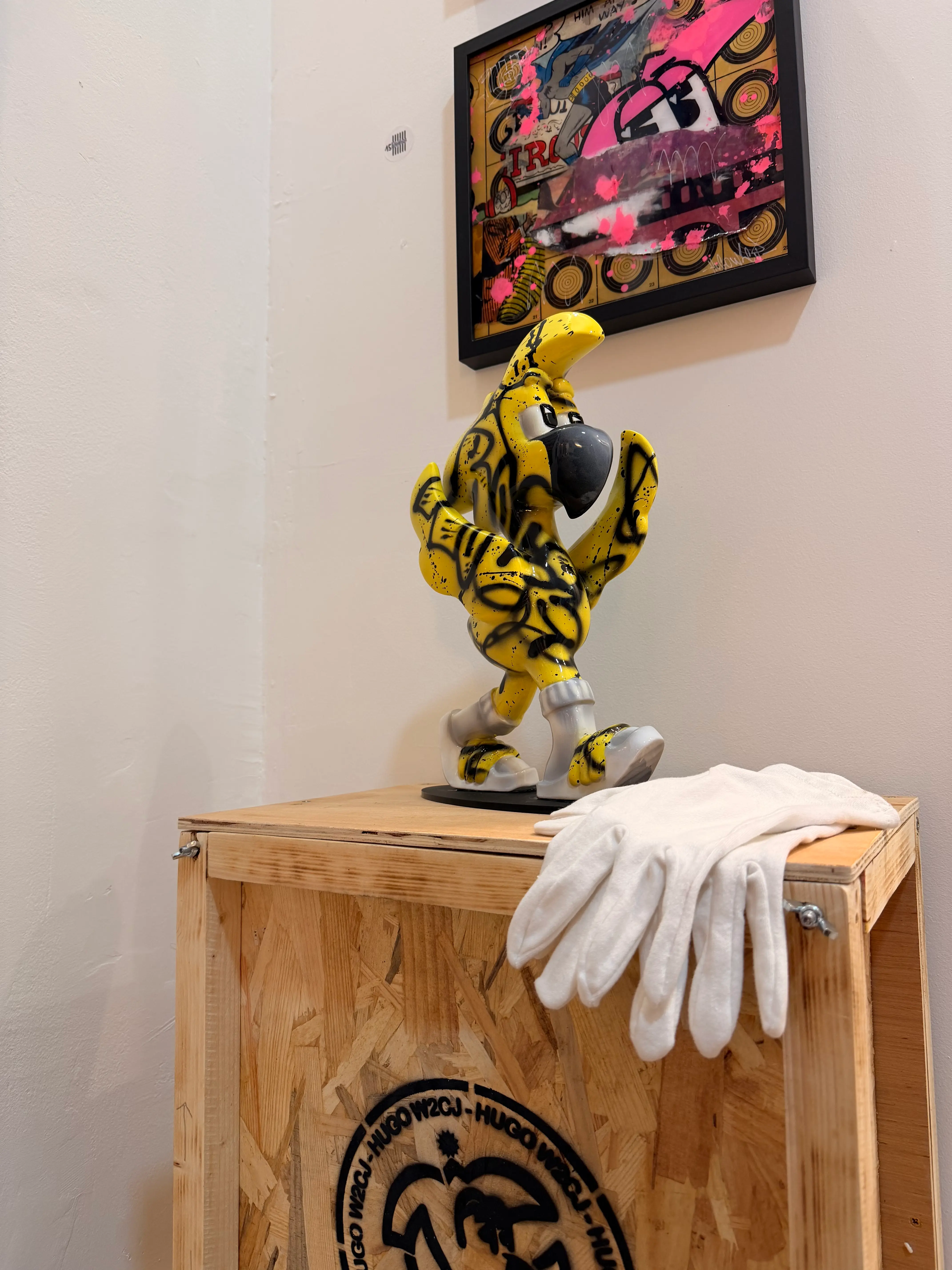 Sculpture de Roger le Flamant Rose jaune graffée de noir, posée sur une caisse en bois marquée Hugo W2CJ, avec des gants blancs