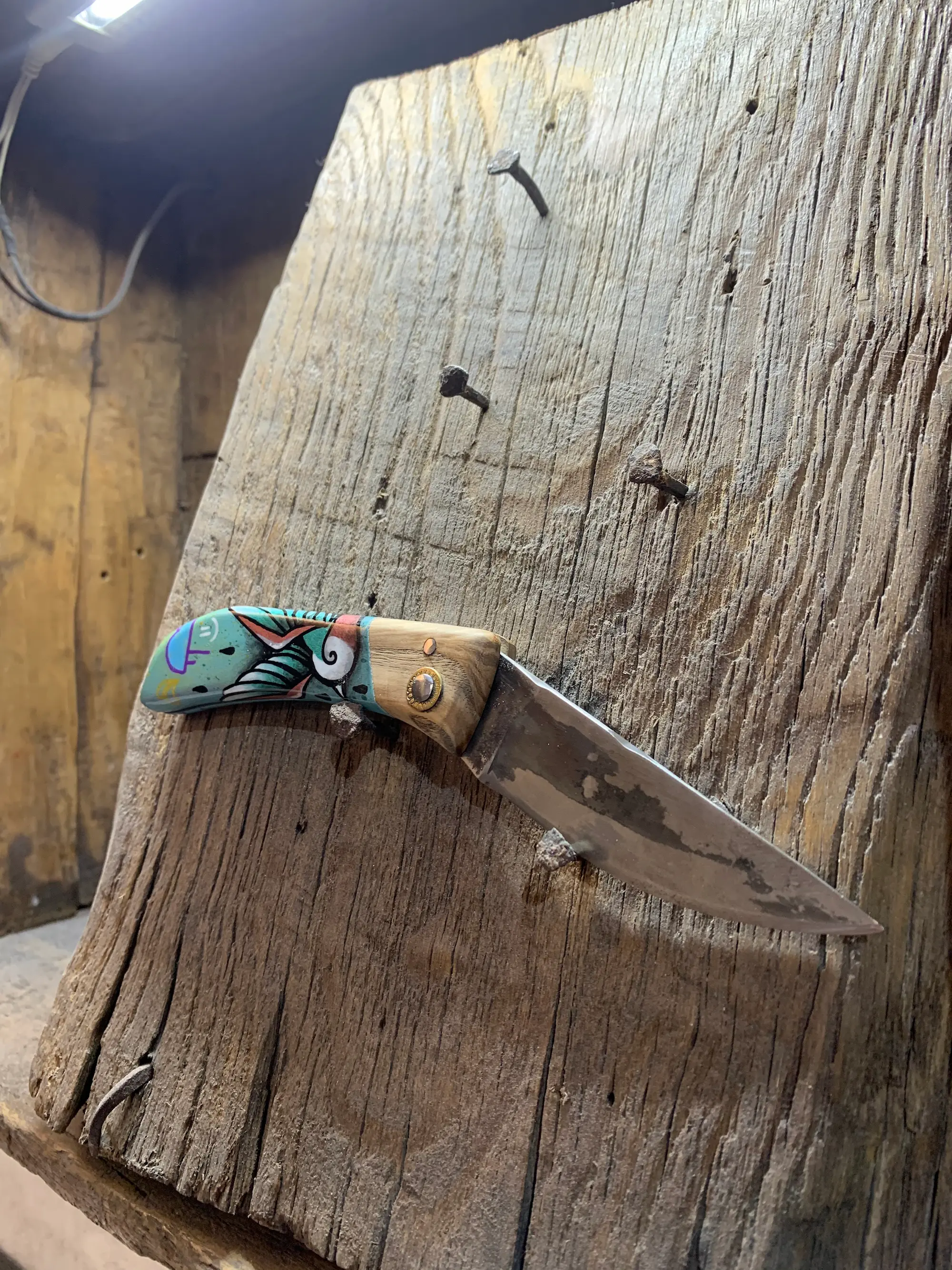 Couteau pliant de Manu Padovani au manche peint par Hugo W2CJ de motifs graphiques turquoise, rouges et blancs, posé sur une poutre en bois