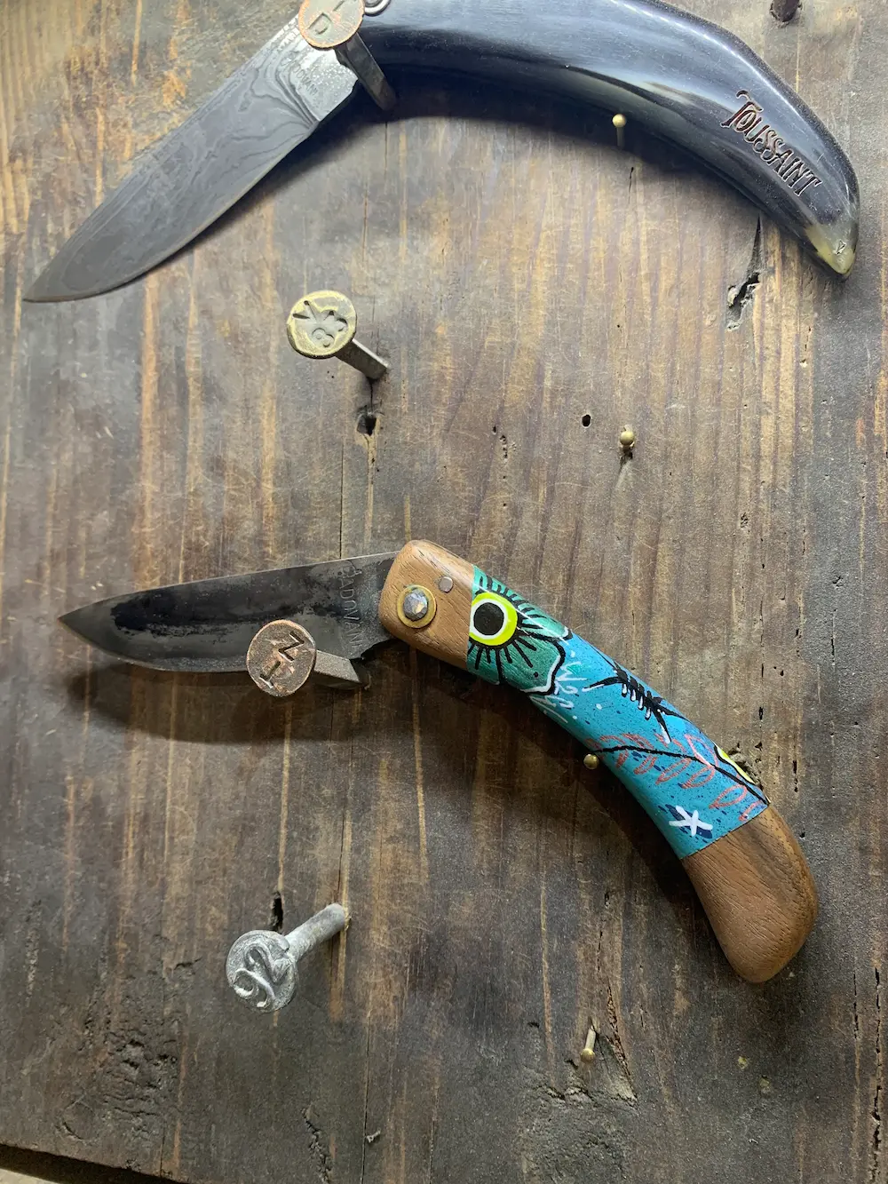Couteau pliant de Manu Padovani au manche peint par Hugo W2CJ en turquoise, avec un œil et des branches, accroché sur une planche de bois