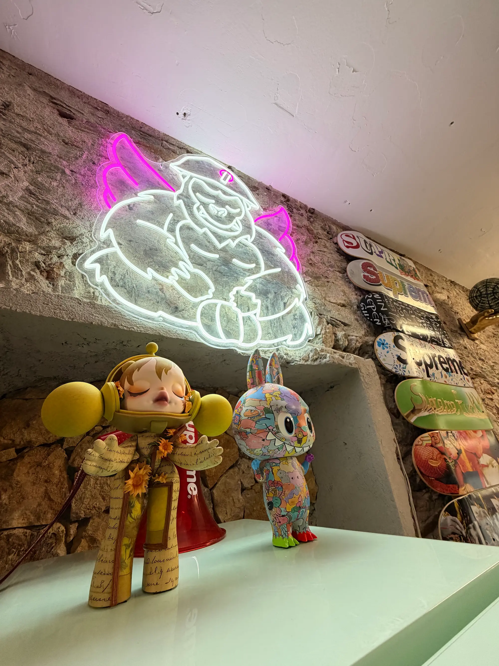 Néon blanc et rose reprenant la mascotte ailée de Chez Jean dessinée par Hugo W2CJ, au-dessus de figurines de collection