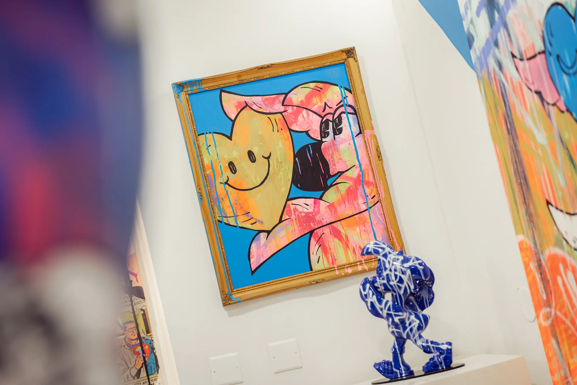 Toile au cadre doré où Roger le Flamant Rose serre un cœur jaune souriant, à côté d'une sculpture bleue