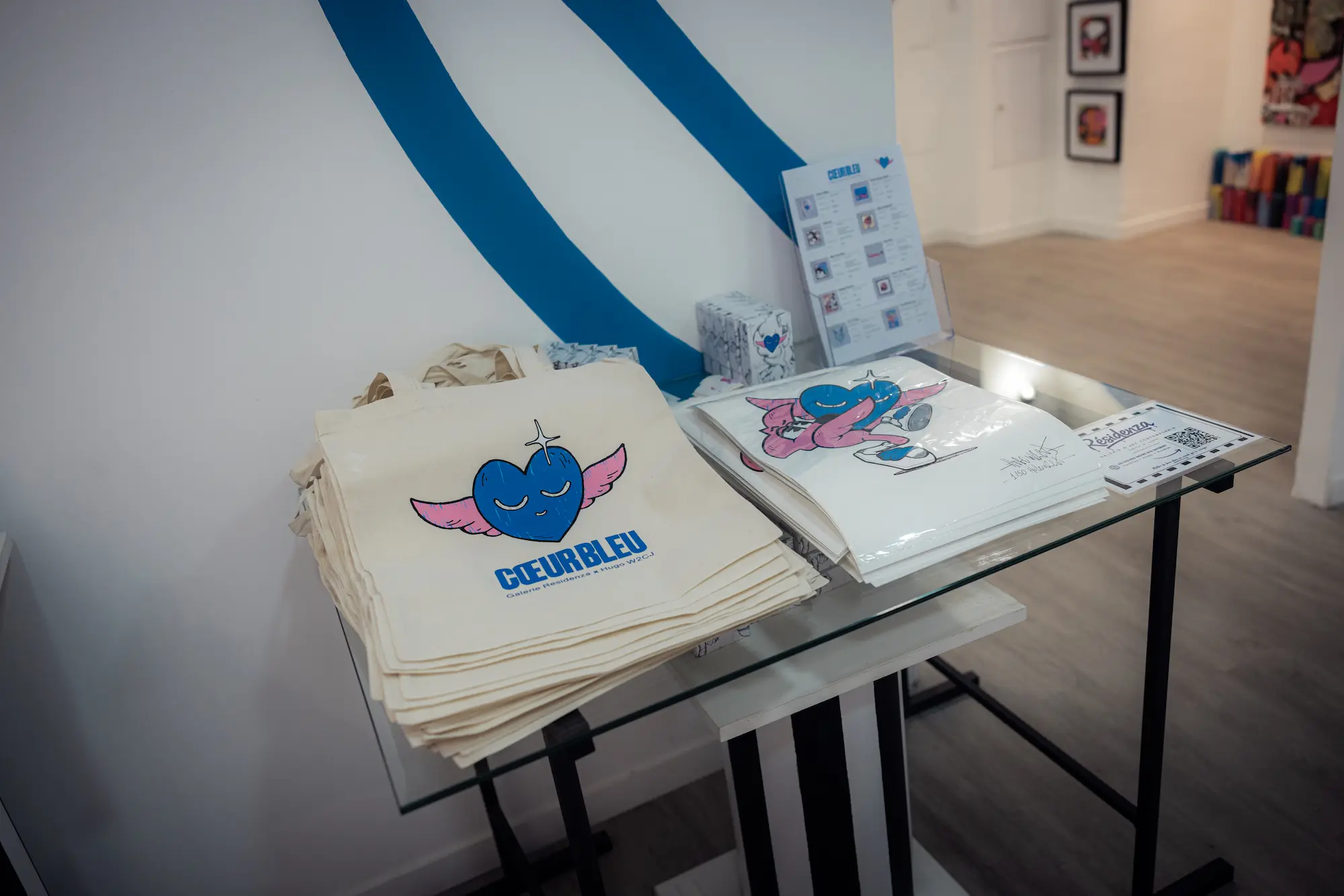 Tote bags Cœur Bleu et catalogue posés sur une table en verre dans la galerie