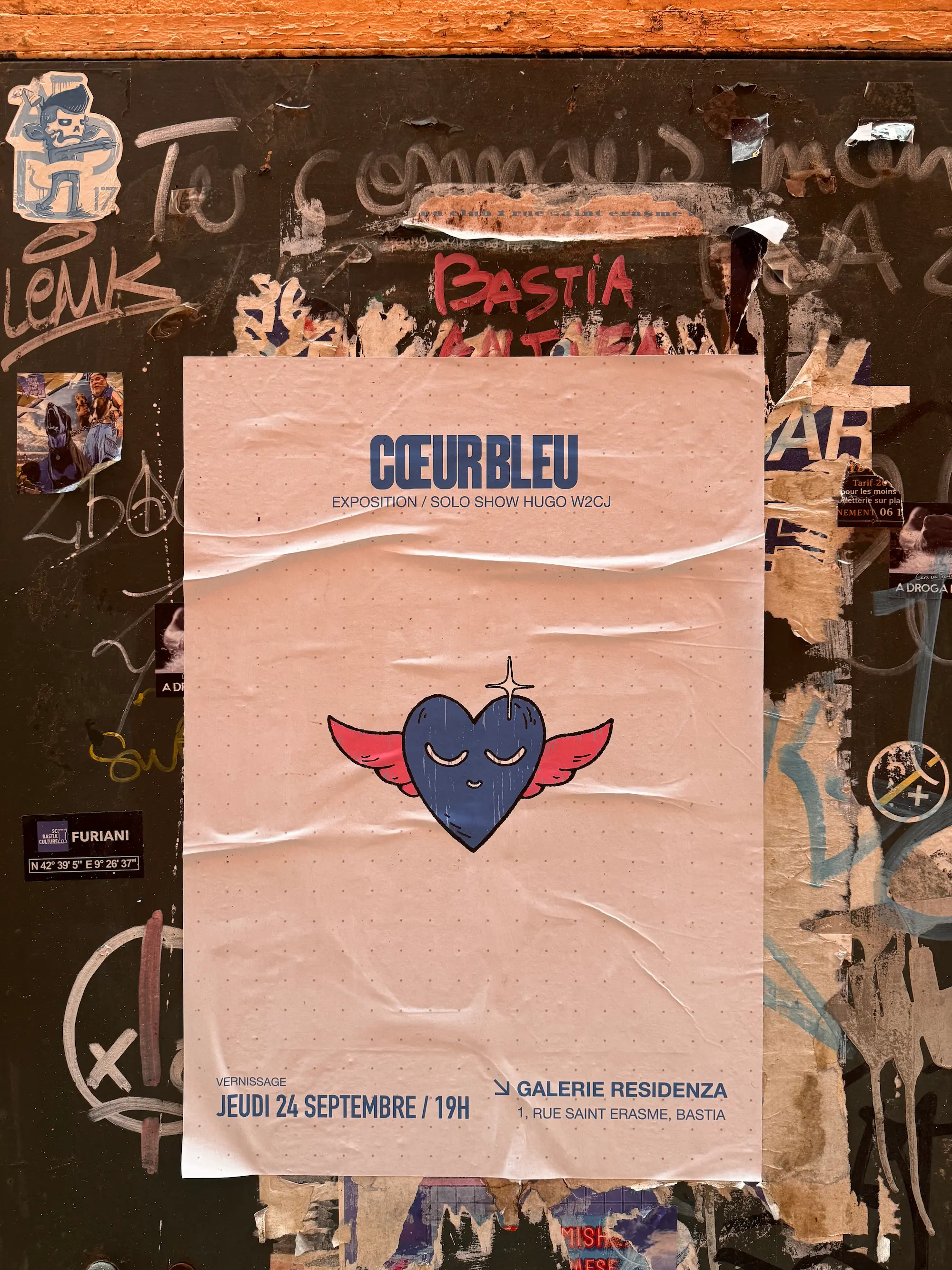 Affiche de l'exposition Cœur Bleu collée sur un mur couvert de graffitis, dans une rue de Bastia