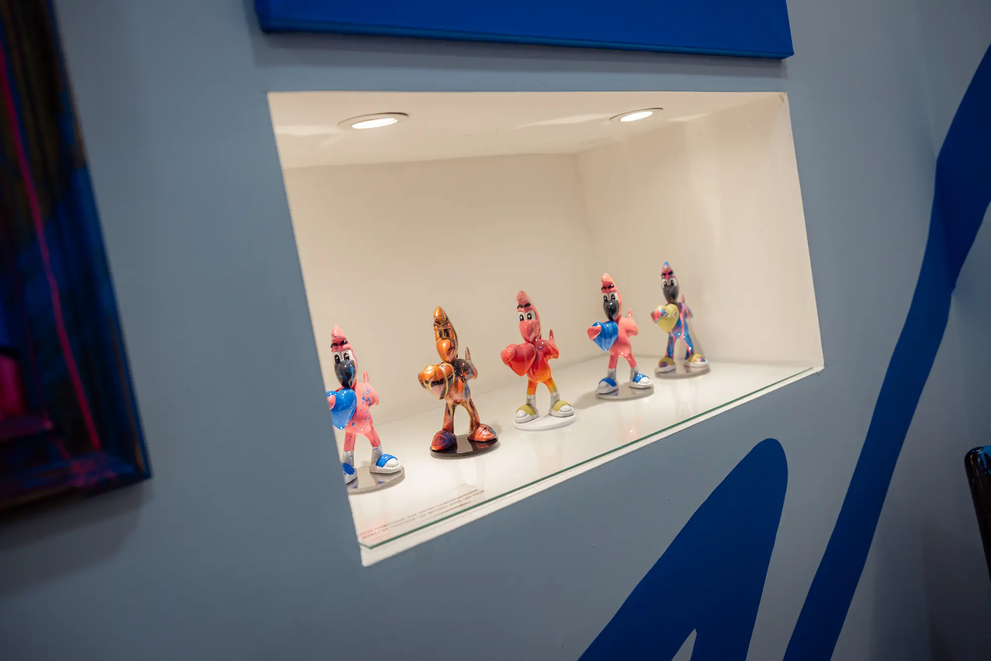 Niche éclairée où sont alignées cinq petites sculptures de Roger le Flamant Rose tenant un cœur, série Heart Delivery, exposition Cœur Bleu