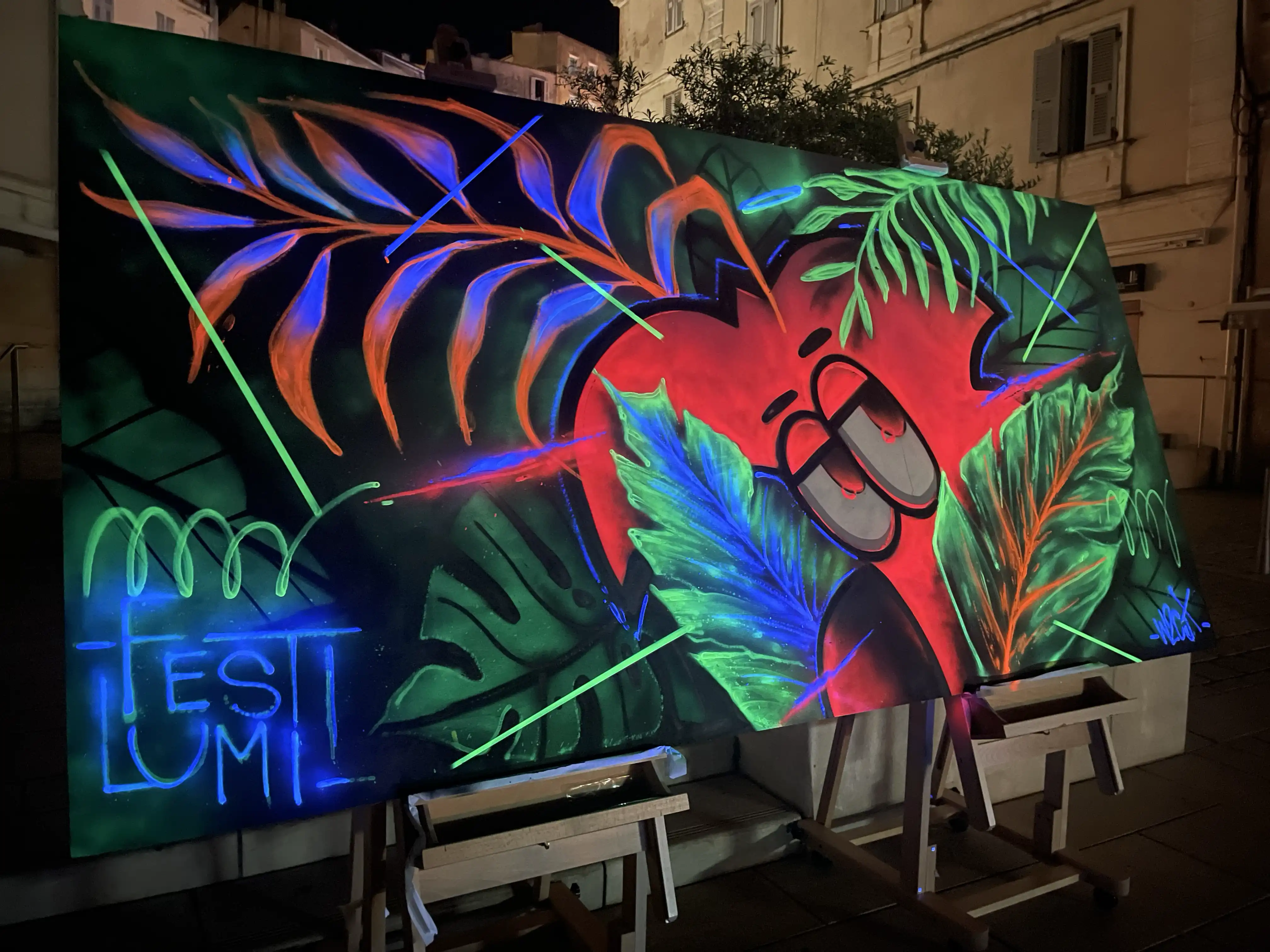 Live painting au festival Festi Lumi, Bonifacio, 2022 : grande toile fluorescente sous lumière noire, Roger le Flamant Rose parmi des feuilles tropicales, de nuit sur une place