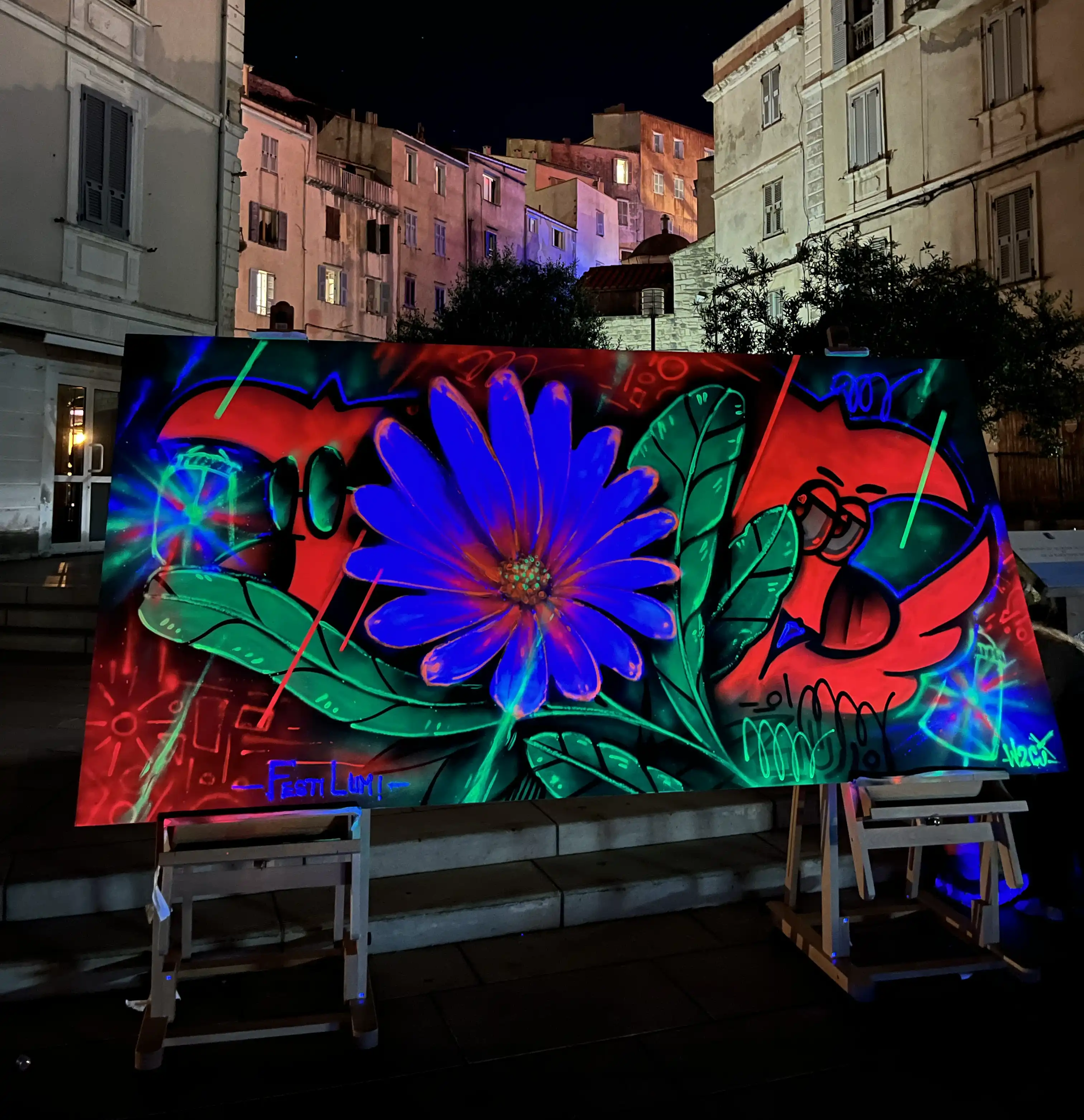 Live painting au festival Festi Lumi, Bonifacio, 2022 : toile fluorescente sous lumière noire, une fleur bleue géante et Roger le Flamant Rose, devant les façades de la vieille ville de nuit