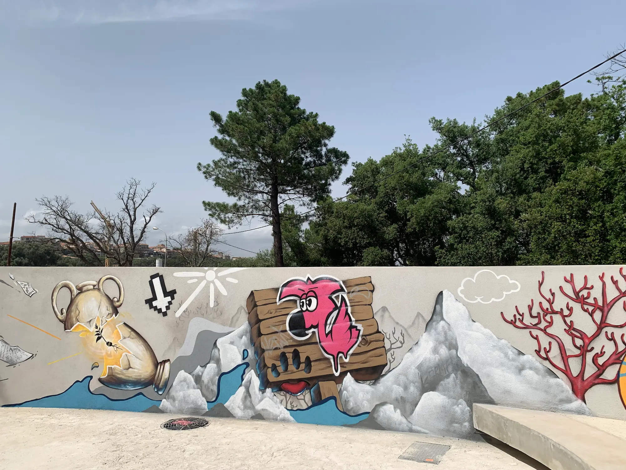 Fresque du jardin de la médiathèque L'animu, avec Piombu, 2021 : Roger le Flamant Rose sur une caisse en bois, amphore, montagnes et corail rouge sur un long mur